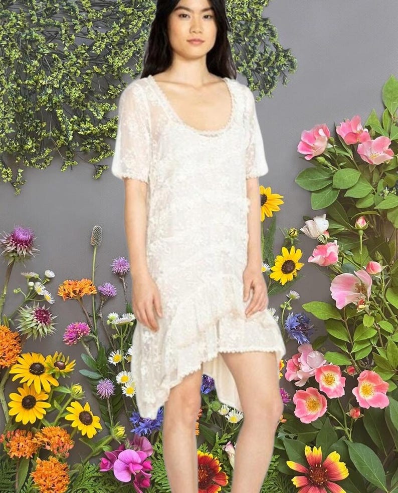 May include: A white lace dress with a scoop neckline and short sleeves. The dress has a layered, asymmetrical hemline. The background features a gray wall adorned with colorful flowers and greenery.