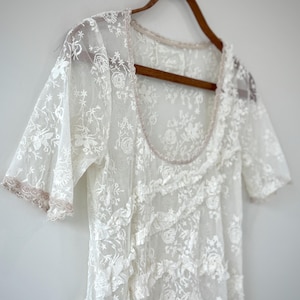 May include: A white lace dress with short sleeves and a scoop neckline hangs on a wooden hanger. The dress features floral embroidery and ruffled detailing. The edges of the sleeves and neckline are trimmed with a light pink trim.
