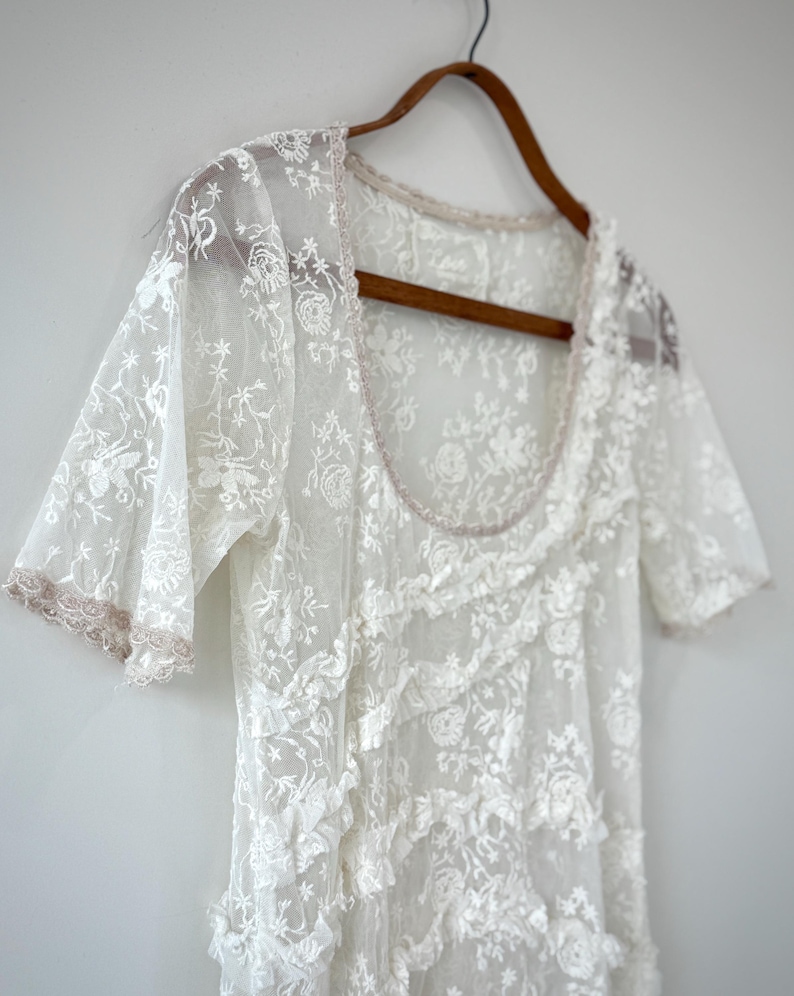 May include: A white lace dress with short sleeves and a scoop neckline hangs on a wooden hanger. The dress features floral embroidery and ruffled detailing. The edges of the sleeves and neckline are trimmed with a light pink trim.