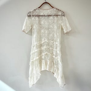 May include: A vintage-style, ivory lace dress with short sleeves and a tiered, asymmetrical hemline. The dress is adorned with floral patterns and delicate ruffles, hanging on a wooden hanger against a plain white background. The dress is a delicate, romantic piece.