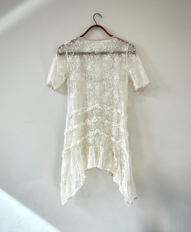 May include: A vintage-style, ivory lace dress with short sleeves and a tiered, asymmetrical hemline. The dress is adorned with floral patterns and delicate ruffles, hanging on a wooden hanger against a plain white background. The dress is a delicate, romantic piece.