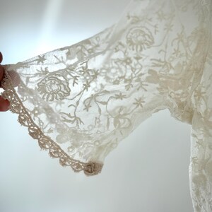 May include: Close-up of a cream-colored lace sleeve. The delicate fabric features floral and star patterns, with a scalloped edge. The sleeve is partially visible, showcasing the intricate details of the lace.