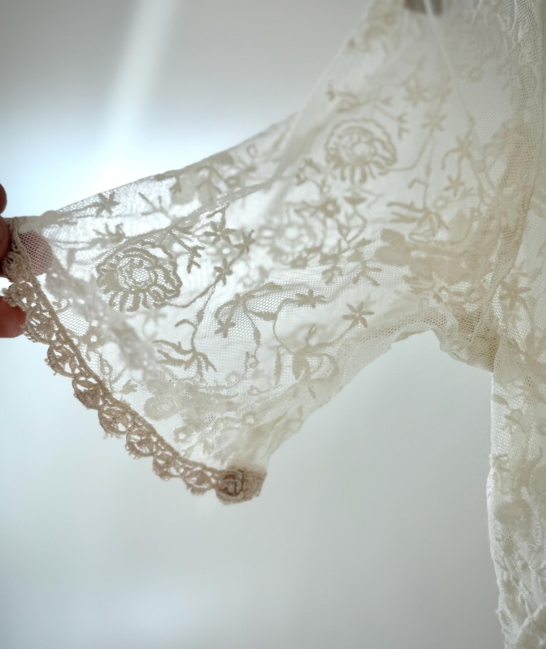 May include: Close-up of a cream-colored lace sleeve. The delicate fabric features floral and star patterns, with a scalloped edge. The sleeve is partially visible, showcasing the intricate details of the lace.