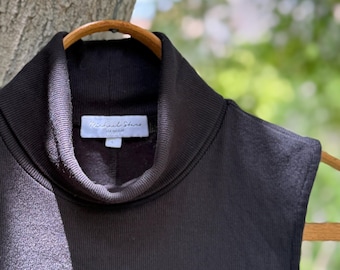 May include: Black turtleneck top with a ribbed texture, hanging on a wooden hanger. The label inside the collar reads "Michael Stars". The background is a blurred view of green foliage.