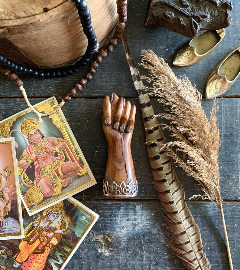 May include: A wooden hand sculpture with a silver filigree base, alongside religious cards, beads, feathers, and brass decorative elements. The items are arranged on a dark wooden surface, creating a rustic and spiritual aesthetic.