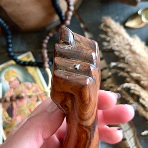 May include: A carved wooden fist sculpture, featuring detailed grain patterns and a polished finish. The sculpture is a warm brown color and appears to be hand-carved. The fist is clenched, with the thumb resting on the index finger.