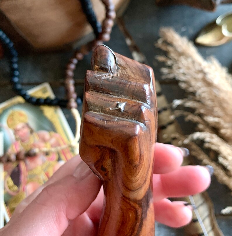 May include: A carved wooden fist sculpture, featuring detailed grain patterns and a polished finish. The sculpture is a warm brown color and appears to be hand-carved. The fist is clenched, with the thumb resting on the index finger.