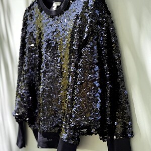 K&ouml;nnte beinhalten: Schwarzes Pailletten-Sweatshirt mit Rundhalsausschnitt und langen &Auml;rmeln. Das Oberteil ist mit gl&auml;nzenden schwarzen Pailletten bedeckt, die einen schimmernden Effekt erzeugen. Die &Auml;rmel und der Ausschnitt sind einfarbig schwarz.