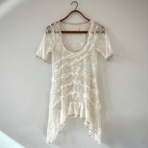 May include: Ivory lace top with short sleeves and a scoop neckline. The top features multiple layers of ruffled lace and a scalloped hemline. It hangs on a wooden hanger against a white wall.