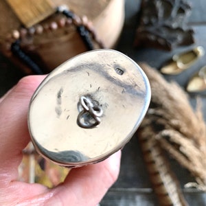 May include: A close-up of a silver-colored, oval-shaped object with a loop. The object has a shiny, reflective surface and a small marking. The background includes a wooden surface and decorative elements.
