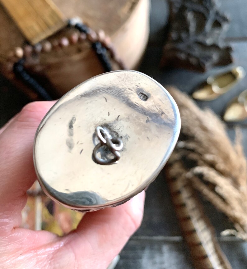 May include: A close-up of a silver-colored, oval-shaped object with a loop. The object has a shiny, reflective surface and a small marking. The background includes a wooden surface and decorative elements.