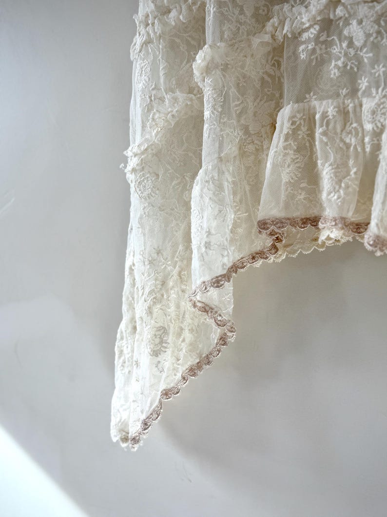 May include: A section of a white lace curtain with multiple layers and floral patterns. The edges are trimmed with a delicate, scalloped, light brown lace. The curtain is hanging against a white wall, with soft lighting.