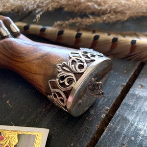 May include: A wooden object with a silver metal band featuring an ornate, openwork design. The band encircles a flat, oval surface. The wood has a warm, brown tone, and the metal has a tarnished appearance.