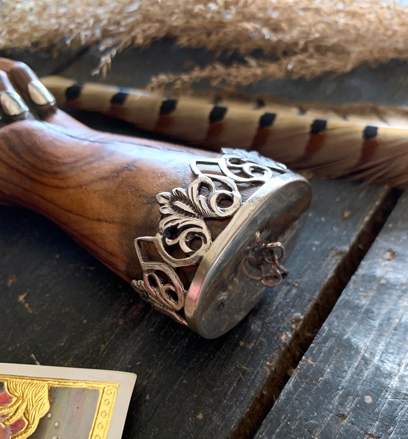 May include: A wooden object with a silver metal band featuring an ornate, openwork design. The band encircles a flat, oval surface. The wood has a warm, brown tone, and the metal has a tarnished appearance.