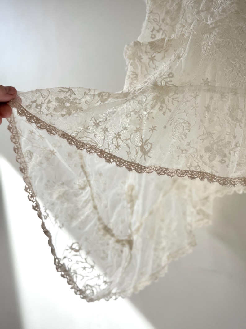 May include: Close-up of a cream-colored lace fabric with intricate floral patterns. The delicate lace features a scalloped edge with a contrasting trim. The fabric is held up, showcasing its texture and design.