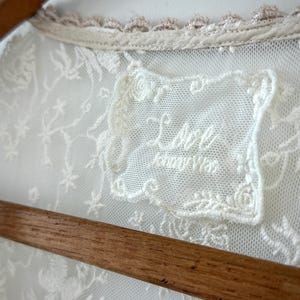 May include: Close-up of a cream-colored lace garment with floral embroidery. The garment features a delicate lace collar with a light brown trim. A label with the text "Lace Honey Was" is visible.