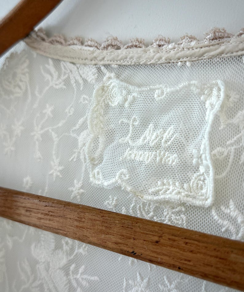 May include: Close-up of a cream-colored lace garment with floral embroidery. The garment features a delicate lace collar with a light brown trim. A label with the text "Lace Honey Was" is visible.