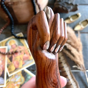 May include: A carved wooden hand in a fist, with silver-colored nail accents. The hand is a warm brown color, with visible wood grain. The background includes religious imagery and decorative items.