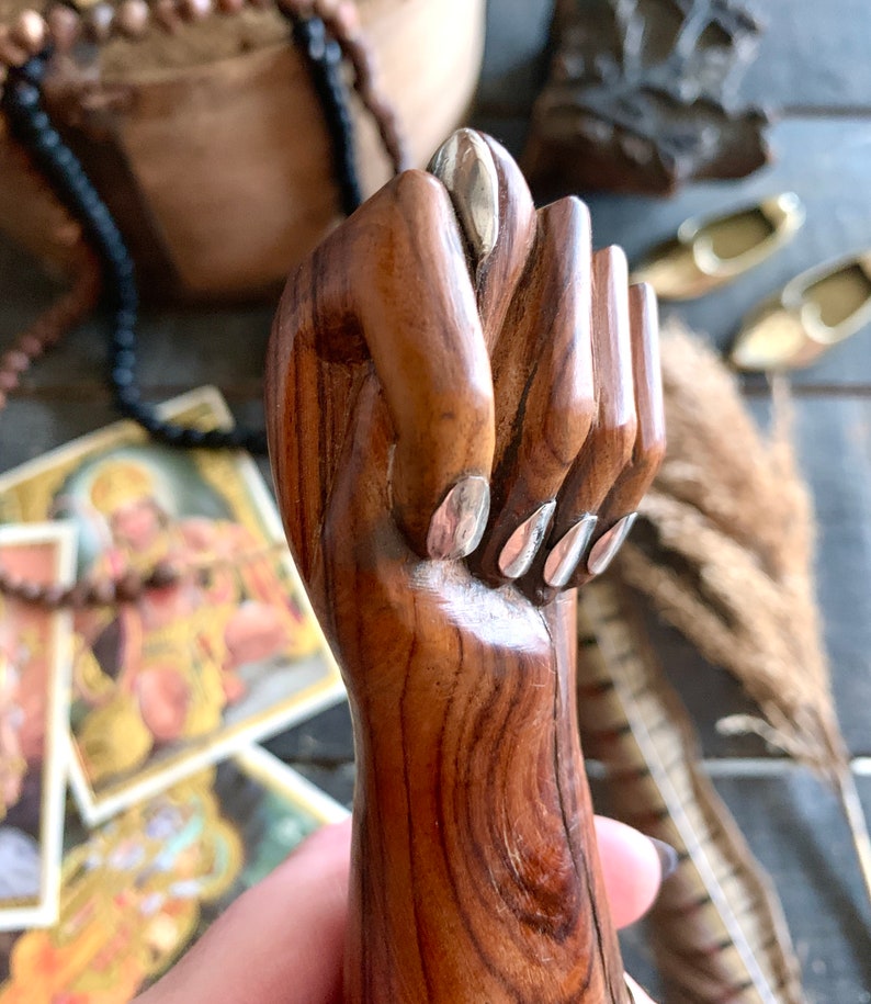 May include: A carved wooden hand in a fist, with silver-colored nail accents. The hand is a warm brown color, with visible wood grain. The background includes religious imagery and decorative items.