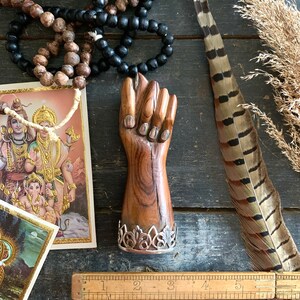 May include: A wooden fist sculpture with a silver filigree base. The sculpture is brown with silver accents. The image also includes prayer beads, a feather, and a vintage ruler.