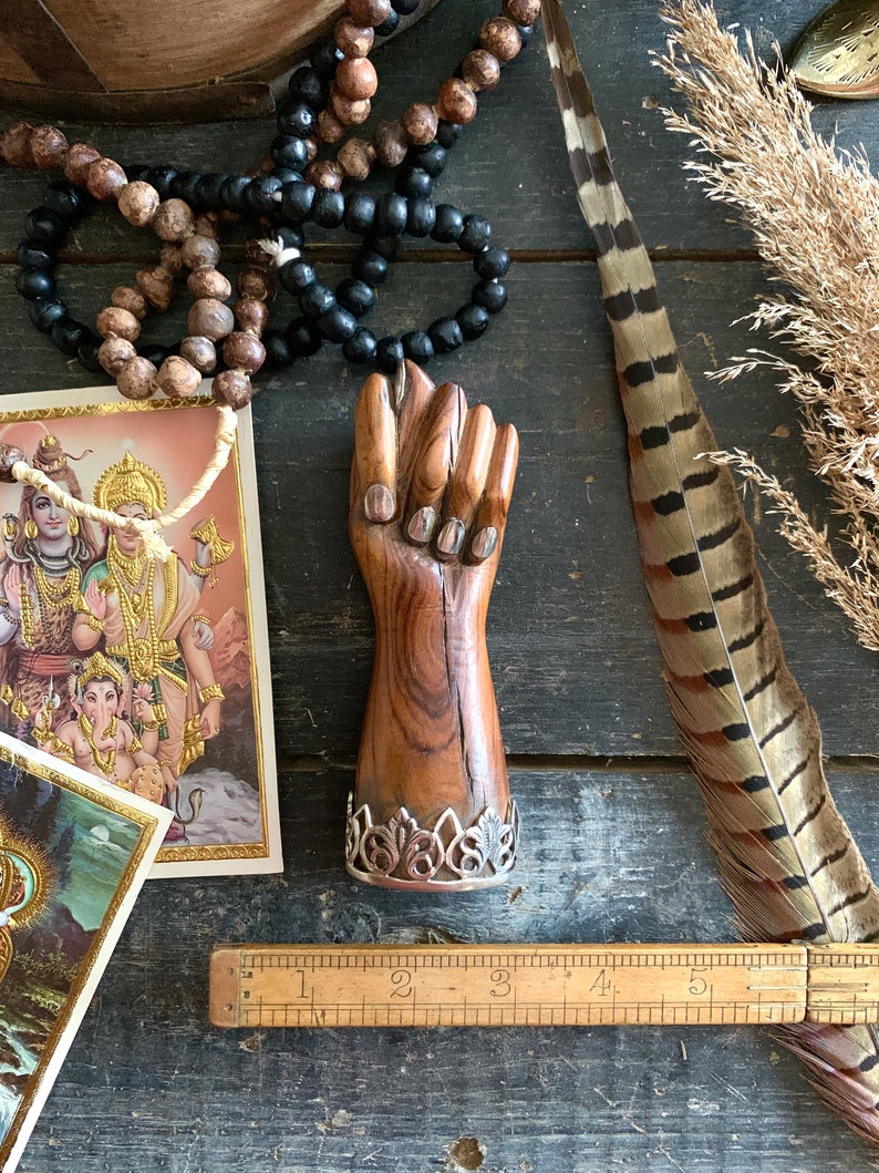 May include: A wooden fist sculpture with a silver filigree base. The sculpture is brown with silver accents. The image also includes prayer beads, a feather, and a vintage ruler.