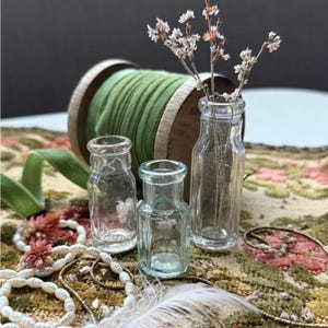 Peut inclure: Trois petites bouteilles en verre transparent de formes variées, dont une contenant des fleurs séchées. Une bobine en bois avec un ruban vert et un fil de perles sont en arrière-plan. Les objets sont disposés sur un tissu à motifs floraux.