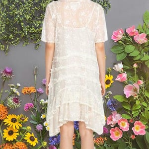 May include: A cream-colored, short-sleeved dress with a delicate lace overlay. The dress features tiered ruffles and a slightly asymmetrical hemline. The background is a floral arrangement with pink, yellow, and orange flowers.