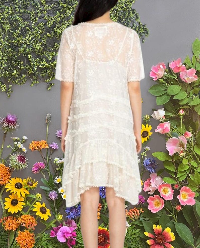 May include: A cream-colored, short-sleeved dress with a delicate lace overlay. The dress features tiered ruffles and a slightly asymmetrical hemline. The background is a floral arrangement with pink, yellow, and orange flowers.