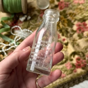 May include: A small, clear glass bottle with embossed text. The bottle reads "Edison Battery Oil Made in U.S.A." and "Thomas A. Edison Incorporated Bloomfield, N.J. U.S.A." The bottle is held in a hand, with a blurred background.