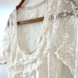 May include: A white lace top with short sleeves and a scoop neckline hangs on a wooden hanger. The delicate fabric features floral embroidery and ruffled details. The garment is a vintage-inspired piece.