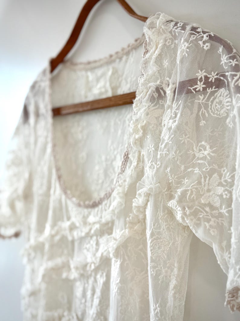 May include: A white lace top with short sleeves and a scoop neckline hangs on a wooden hanger. The delicate fabric features floral embroidery and ruffled details. The garment is a vintage-inspired piece.