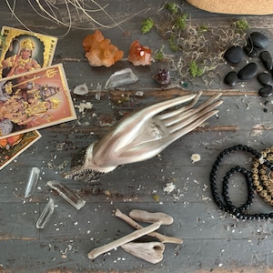May include: A silver-colored hand-shaped decorative object with intricate details, surrounded by crystals, stones, and spiritual cards on a weathered gray wooden surface. The arrangement includes quartz crystals, black beads, and small pieces of driftwood.