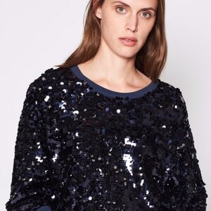 K&ouml;nnte beinhalten: Ein marineblaues Pailletten-Sweatshirt mit einem dunkelblauen Rundhalsausschnitt. Das lang&auml;rmlige Oberteil ist mit gl&auml;nzenden, reflektierenden Pailletten bedeckt. Die Pailletten erzeugen einen strukturierten, schimmernden Effekt und verleihen dem l&auml;ssigen Stil einen Hauch von Glamour.