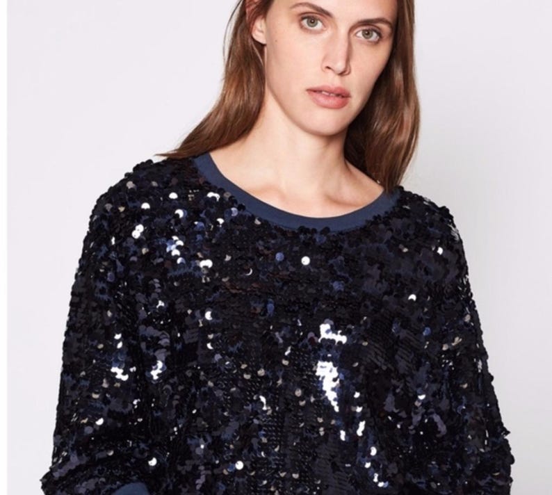 K&ouml;nnte beinhalten: Ein marineblaues Pailletten-Sweatshirt mit einem dunkelblauen Rundhalsausschnitt. Das lang&auml;rmlige Oberteil ist mit gl&auml;nzenden, reflektierenden Pailletten bedeckt. Die Pailletten erzeugen einen strukturierten, schimmernden Effekt und verleihen dem l&auml;ssigen Stil einen Hauch von Glamour.