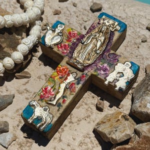 May include: A wooden cross with a gold-colored Virgin Mary figure and silver-colored embellishments. The cross features painted floral designs and blue accents on the edges. A white beaded necklace is visible in the background.