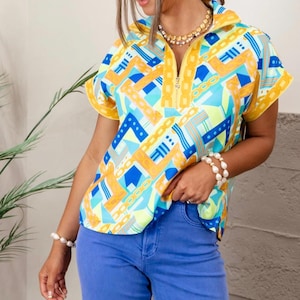 May include: A short-sleeved top with a geometric pattern in yellow, blue, and turquoise. The top has a zipper closure and a yellow collar. The person is wearing blue jeans and gold jewelry. The top's design includes chain and geometric shapes.
