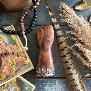May include: A wooden fist-shaped object with a silver filigree base, displayed on a dark wooden surface. The image includes a feather, beads, and religious cards. The fist is a rich brown color, and the silver base adds a decorative touch.