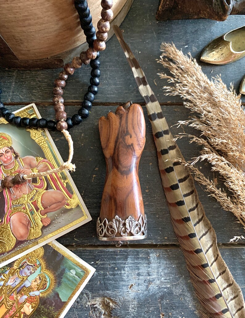 May include: A wooden fist-shaped object with a silver filigree base, displayed on a dark wooden surface. The image includes a feather, beads, and religious cards. The fist is a rich brown color, and the silver base adds a decorative touch.