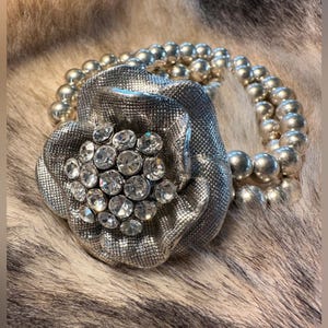 May include: A silver-toned floral brooch with a cluster of clear rhinestones at its center, paired with a beaded necklace. The brooch has a textured, metallic finish, and the necklace features round, silver beads. The jewelry is displayed on a textured, fur-like surface.