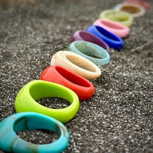 May include: A row of colorful, chunky rings in various shades, including turquoise, green, red, and blue, are arranged on a textured, dark surface. The rings are made of a glossy material and are in focus, showcasing their vibrant colors and smooth surfaces.