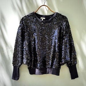 K&ouml;nnte beinhalten: Ein schwarzes Sweatshirt mit Paillettenbesatz, langen &Auml;rmeln und einem Rundhalsausschnitt. Die &Auml;rmel und der Saum sind einfarbig schwarz. Das Kleidungsst&uuml;ck h&auml;ngt an einem goldfarbenen Kleiderb&uuml;gel vor einem hellgr&uuml;nen Hintergrund.