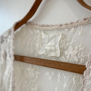 May include: Close-up of a cream-colored lace garment with floral embroidery, hanging on a wooden hanger. The garment has a delicate, scalloped trim in a light pink hue. A small label with text is visible.