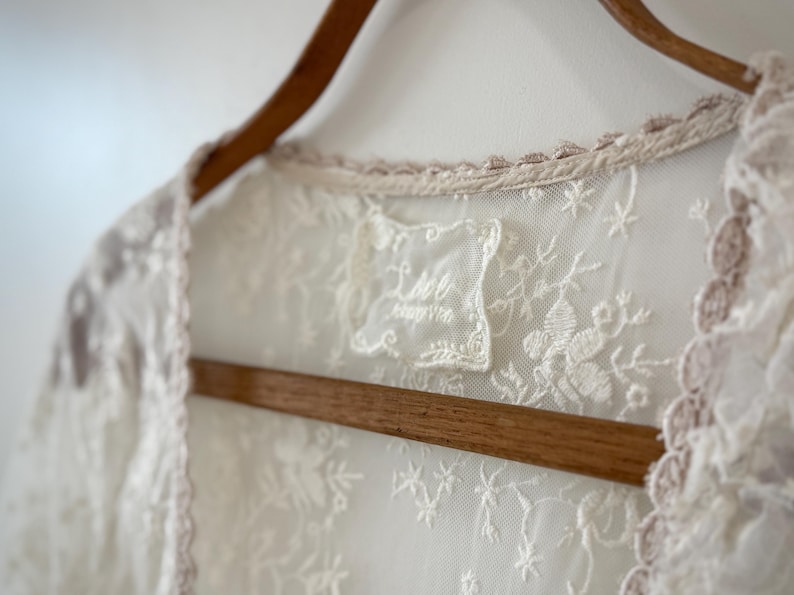 May include: Close-up of a cream-colored lace garment with floral embroidery, hanging on a wooden hanger. The garment has a delicate, scalloped trim in a light pink hue. A small label with text is visible.