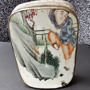 May include: A rectangular, antique porcelain box with a silver filigree lid and gold trim. The box features a hand-painted scene of a person in a blue robe and orange pants in a garden setting. The box has a curved bottom.