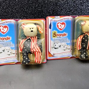 May include: Three Ty Beanie Baby bears in their original packaging. The bears are white with black eyes and noses, and wear outfits with red and white stripes and black with white star patterns. The packaging features the text "Spangle the Bear".