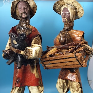 May include: Two handmade paper mache figurines of men in rustic attire. One figure holds a black pig, the other a wooden crate filled with sticks. Both wear hats, red vests, and cream-colored pants. The figures stand on brown bases against a blue backdrop.