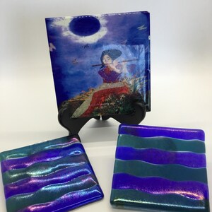 May include: A set of iridescent glass coasters and a decorative panel. The panel features an image of a person playing a flute under a moonlit sky. The coasters have a wavy pattern in shades of blue, purple, and teal.