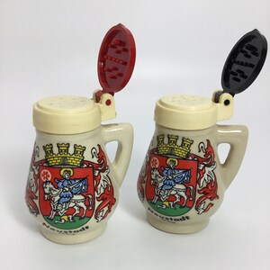 May include: Two ceramic salt and pepper shakers shaped like miniature beer steins. Each stein features a cream-colored body, a handle, and a hinged lid. The lids are red and black. The steins are decorated with a coat of arms and the word "Neustadt".