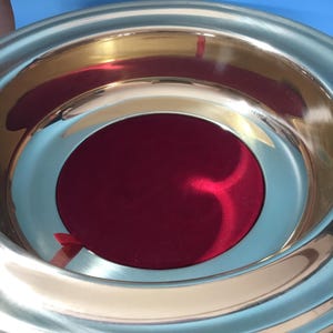 May include: A shiny, silver-colored metal bowl with a deep red, circular interior. The bowl has a wide, reflective rim and a smooth, polished surface. The interior appears to be lined with a soft, velvety material. The bowl is set against a blue background.