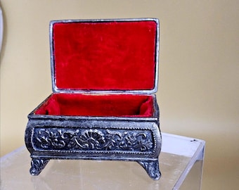 May include: An open jewellery box in an antique style, with a silver-coloured exterior and a vibrant red velvet interior. The box features ornate floral detailing and rests on four small, curved feet. The lid is open, revealing the plush interior.